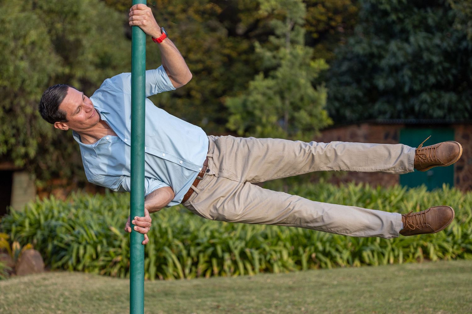groom to be playfully hanging horizontally from a green pole in a lush garden setting.