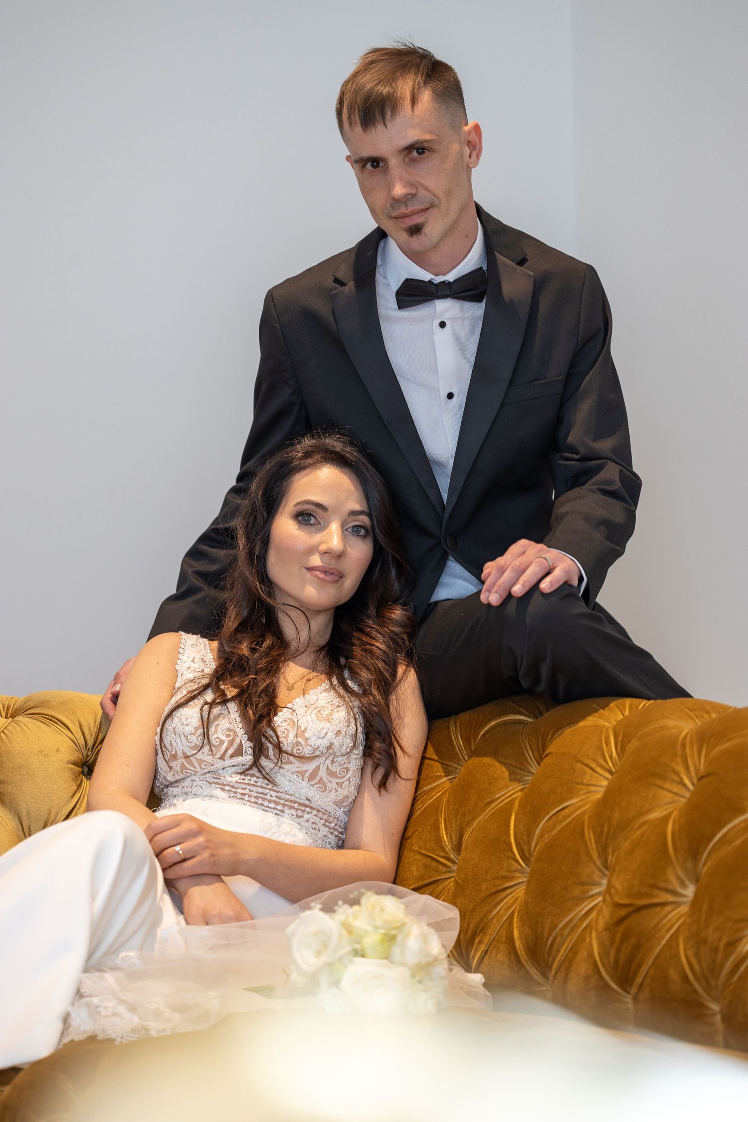 Bride in lace wedding dress reclining on gold velvet sofa with groom standing behind her.