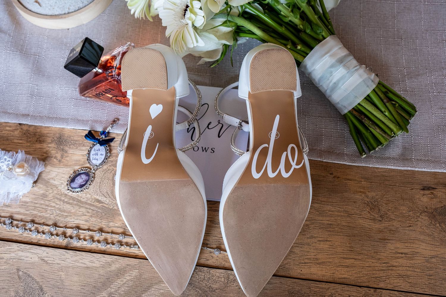Bridal wedding shoes with I Do written on soles displayed with bouquet and accessories on wooden table.