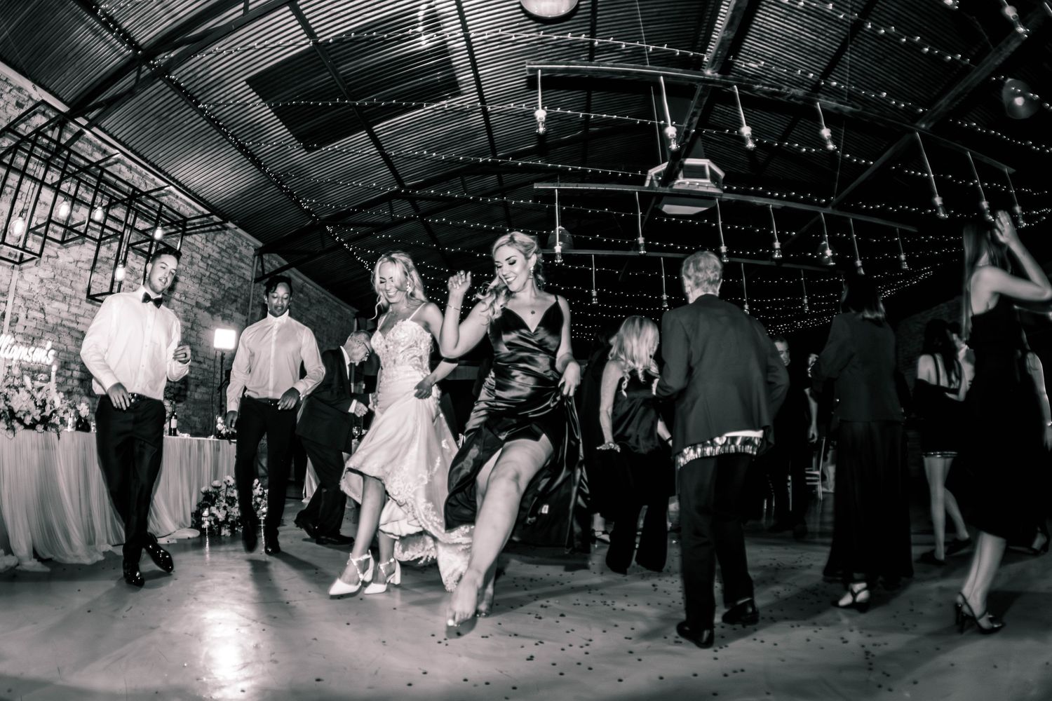 Black and white photo of guests dancing energetically at a rustic wedding reception venue.