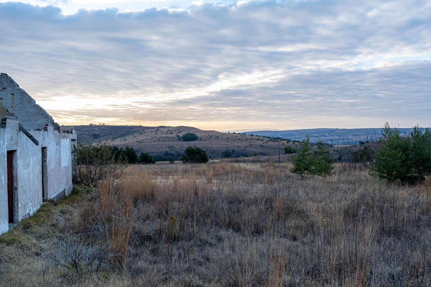 Expansive rural landscape at dusk with dry grassland, distant hills, and dramatic cloudy sky near abandoned building.