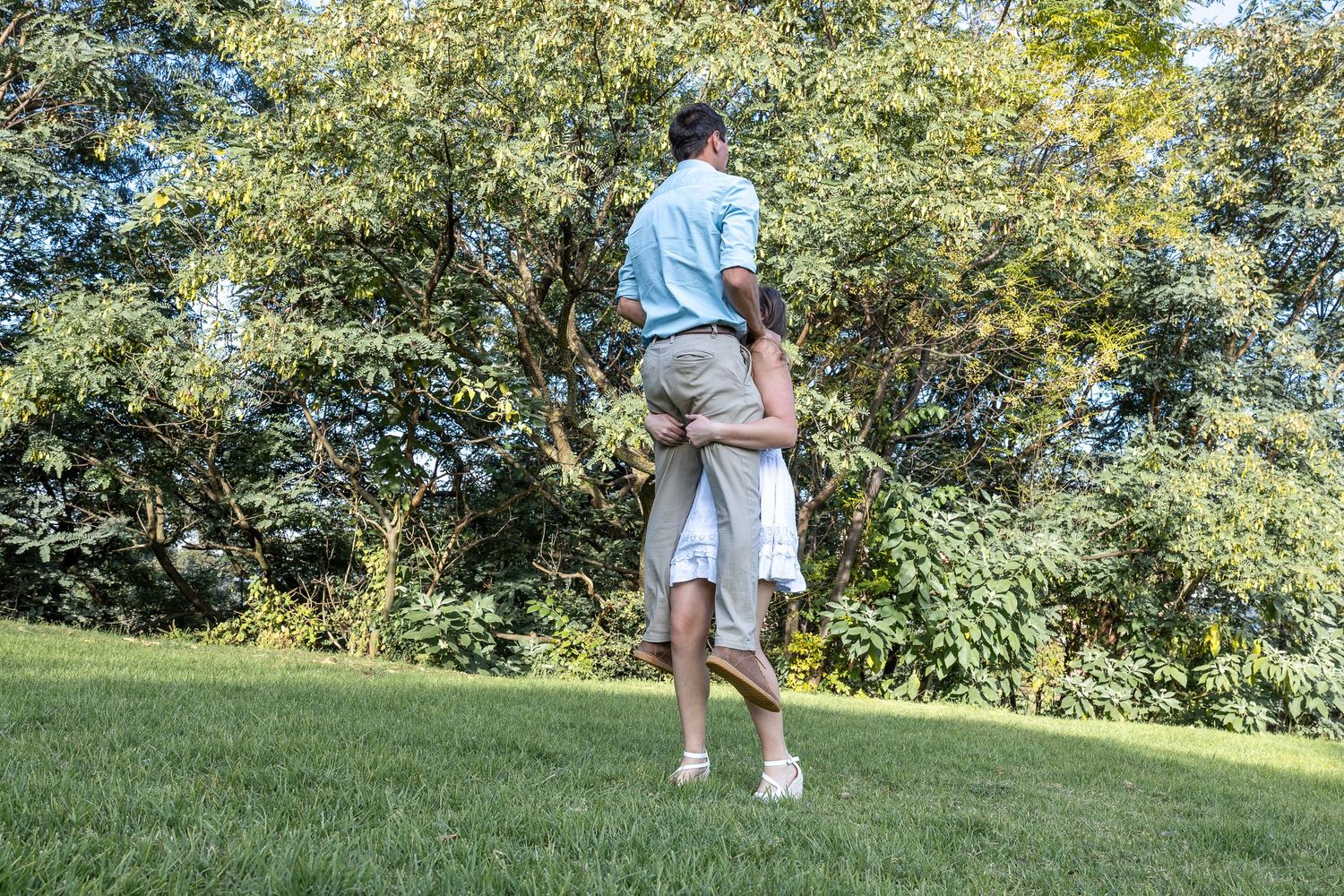 Couple walking hand in hand across green lawn toward lush trees in a garden.
