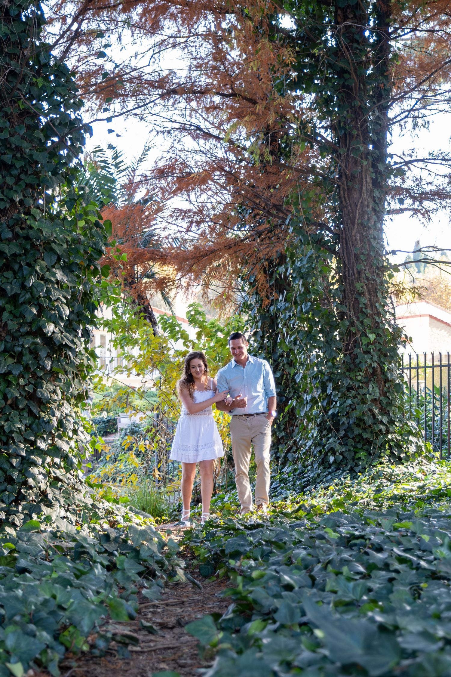 Smiling couple walking hand in hand through a lush ivy-covered garden archway on a sunny day.