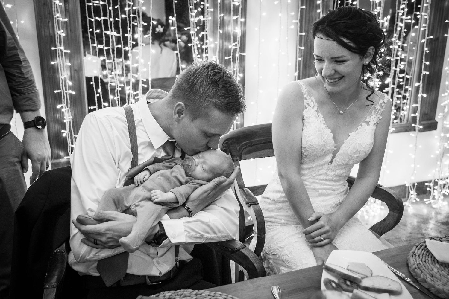 Groom tenderly holds a baby while smiling bride looks on during a heartfelt black and white wedding moment.