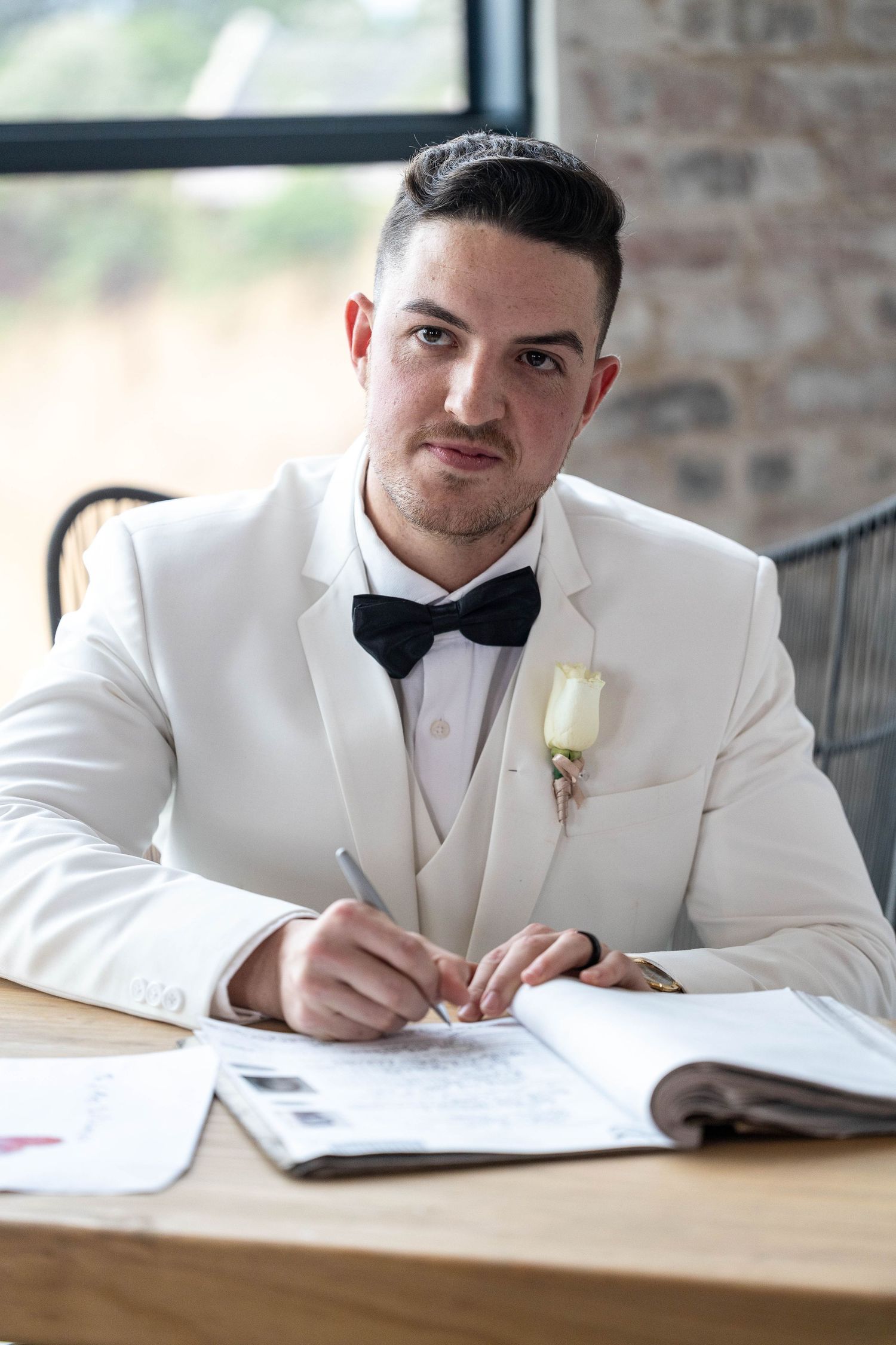 Groom in white tuxedo with black bow tie signs wedding register at wooden table.