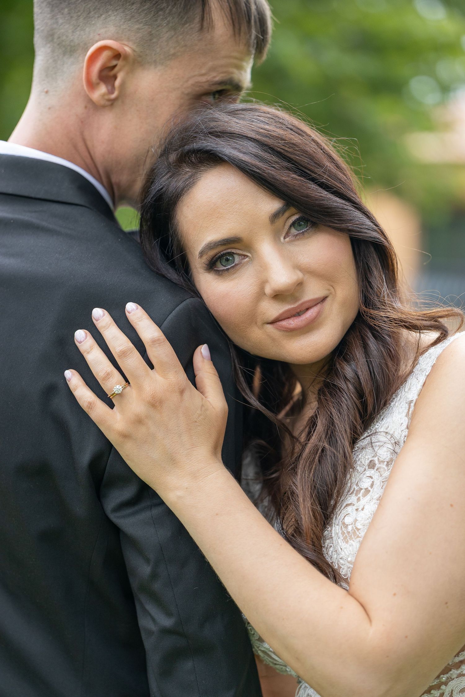 Beautiful brunette bride showing diamond engagement ring while embracing groom in black tuxedo outdoors.