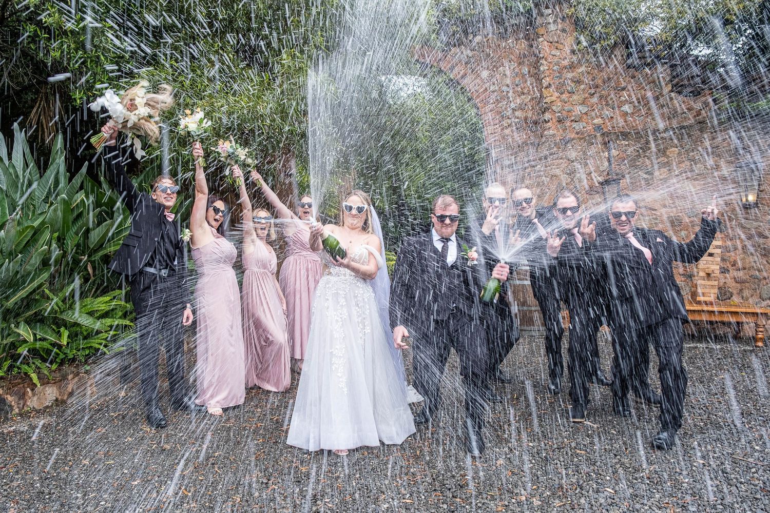 Wedding party celebrates with champagne spray outdoors, bridesmaids in pink dresses holding bouquets.