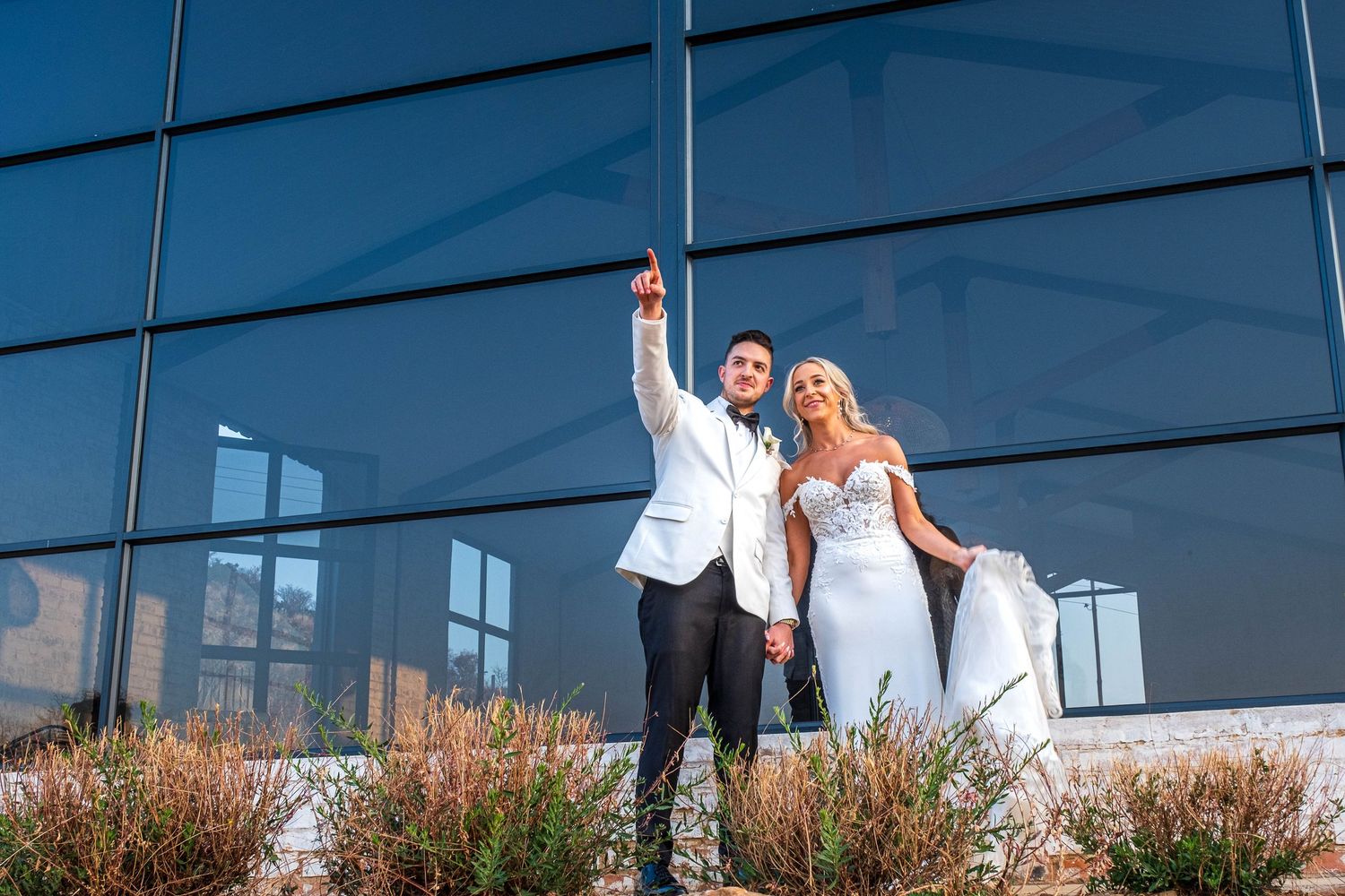 Bride and groom pose together pointing skyward in front of sleek blue glass building on wedding day.