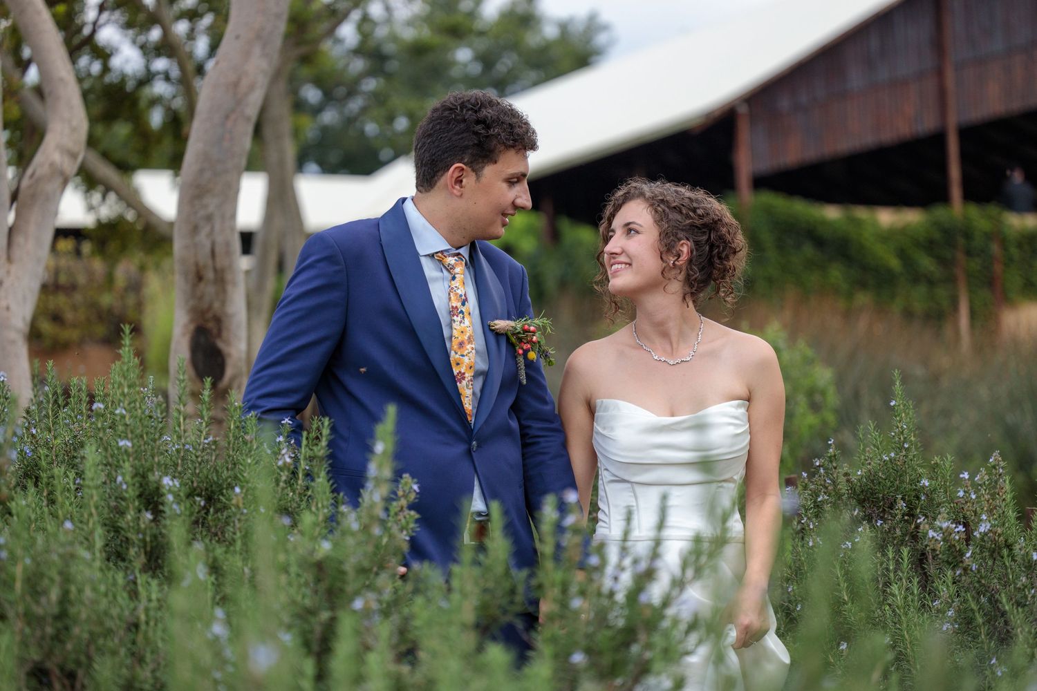 Bride and groom smiling at each other in a field of lavender, groom wearing a blue suit with floral tie.