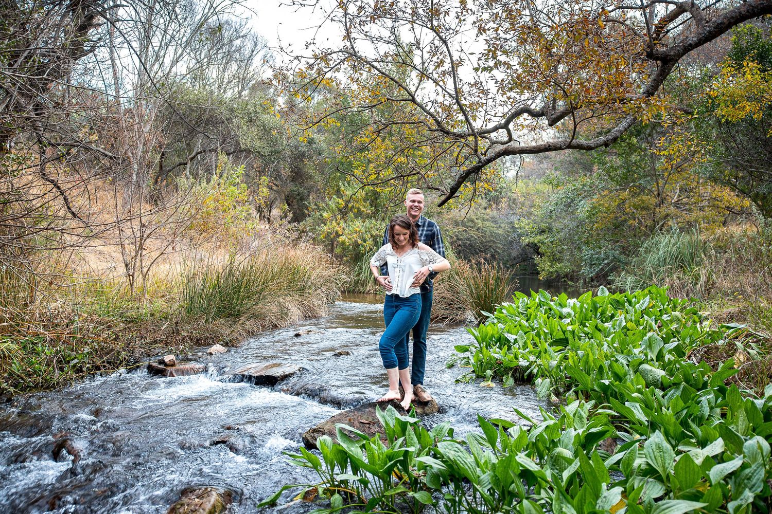 Happy couple standing on rocks in a scenic woodland stream surrounded by lush green foliage.