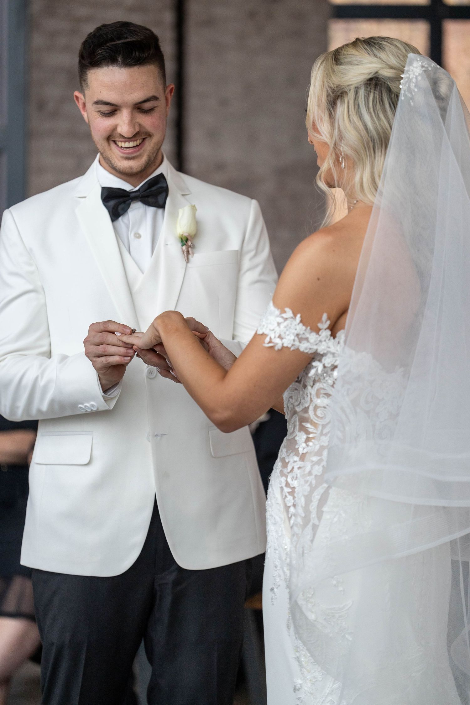 Smiling groom in white tuxedo places wedding ring on brides finger during ceremony.