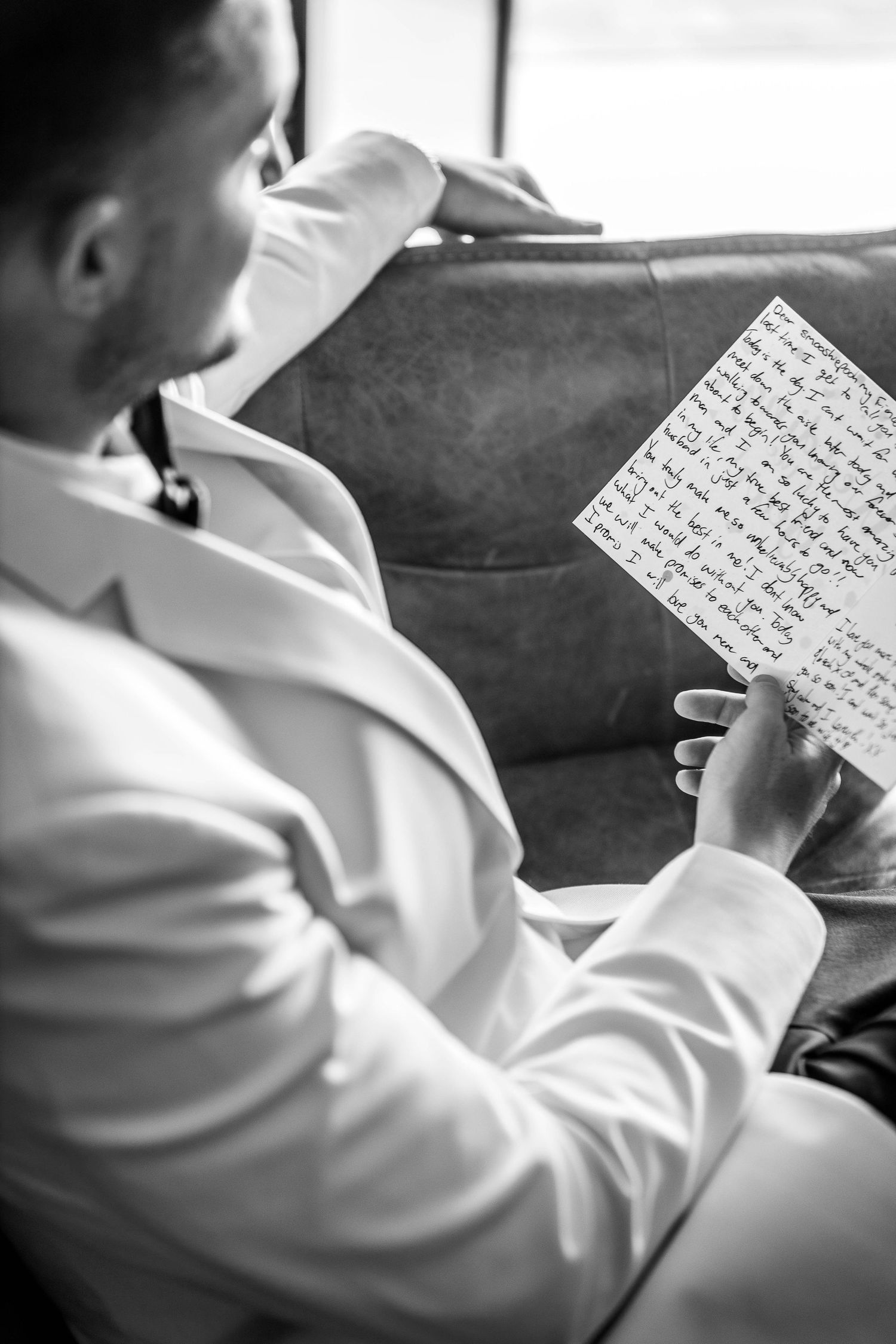 Black and white image of groom reading handwritten letter on leather sofa.