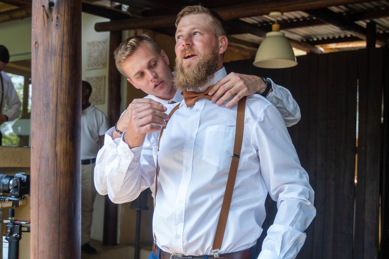 A groomsman adjusts a bow tie on the bearded groom wearing white shirt and brown suspenders before the wedding.