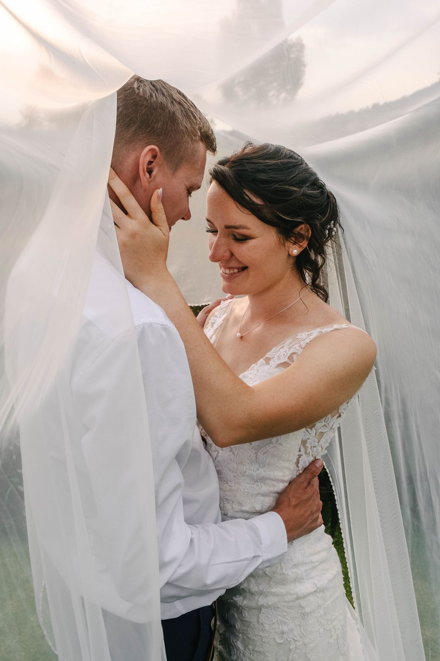 Smiling bride and groom share a tender moment together under a flowing white veil in warm glowing light.