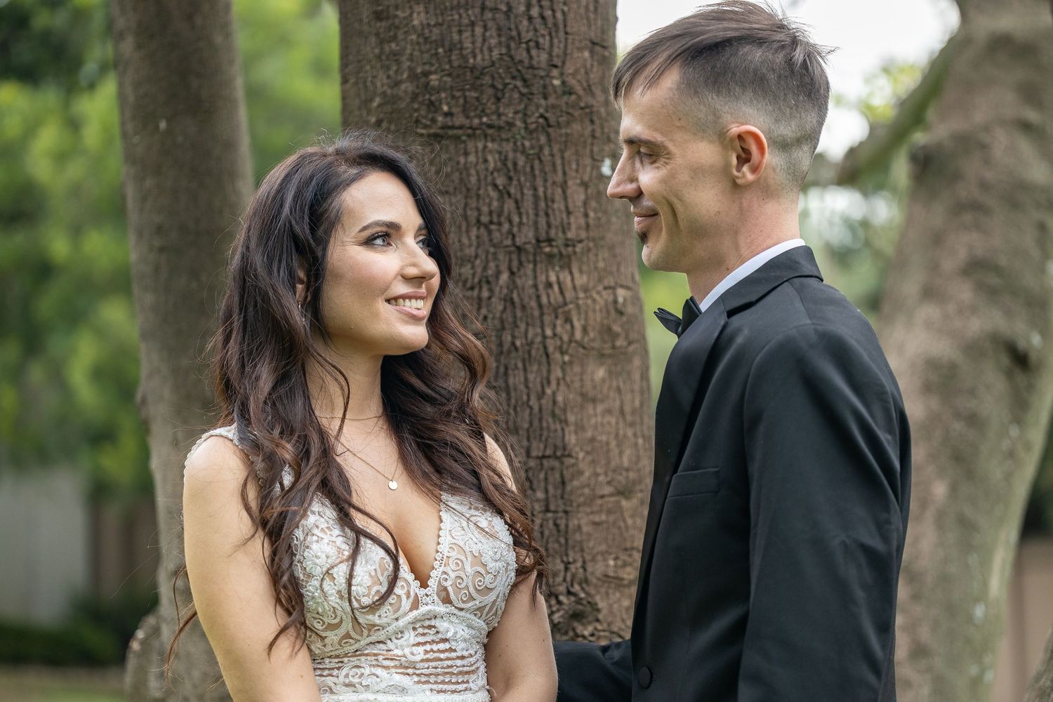 A smiling bride in a lace wedding dress and groom in a black tuxedo share a romantic moment beside a tree.