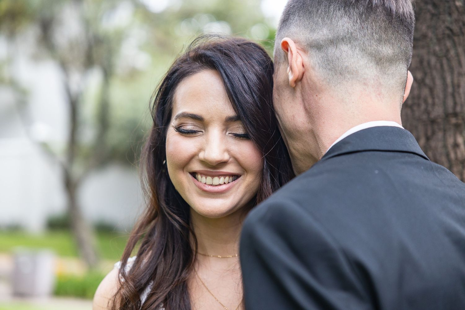 A radiant bride with long dark hair smiles joyfully while leaning close to her groom outdoors.