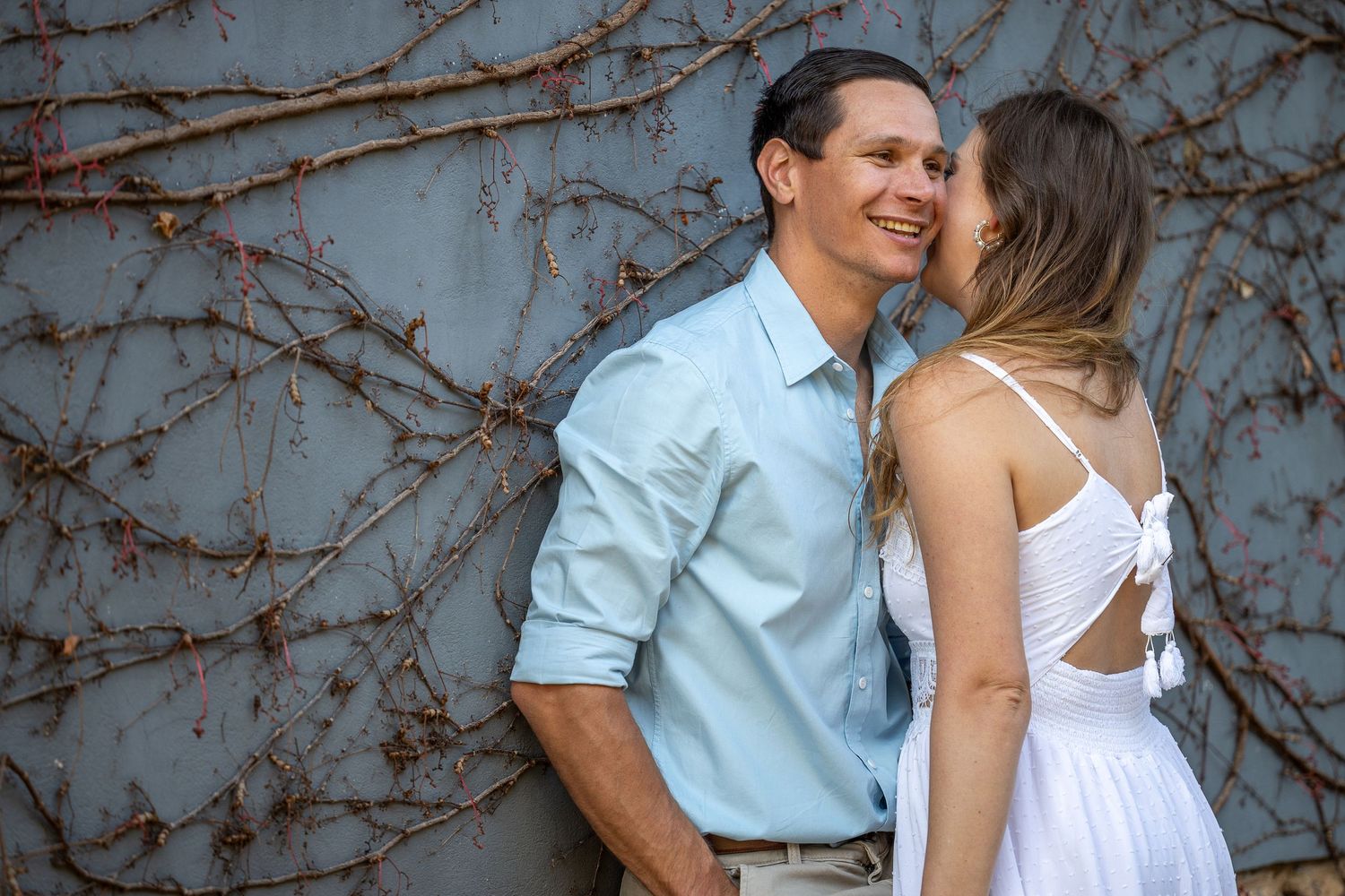 Romantic couple sharing a kiss outdoors against a grey wall with climbing vines.