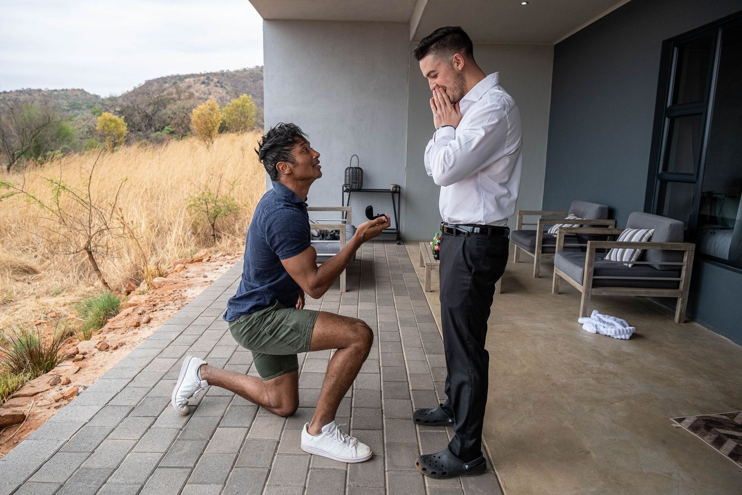 Person proposing on one knee to surprised partner on outdoor terrace.