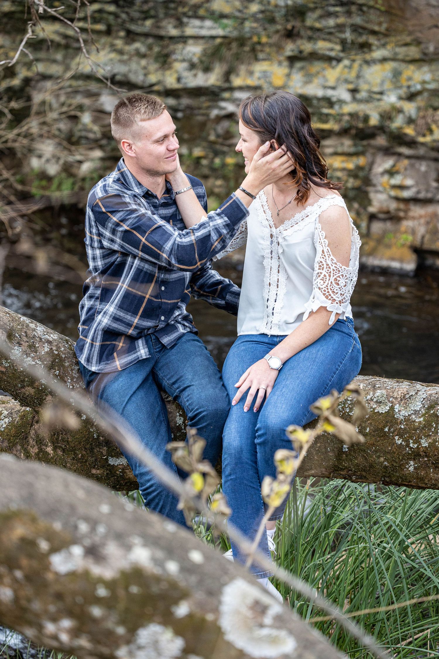 Couple sits on rustic stone fence near creek, tenderly holding each other's faces outdoors.