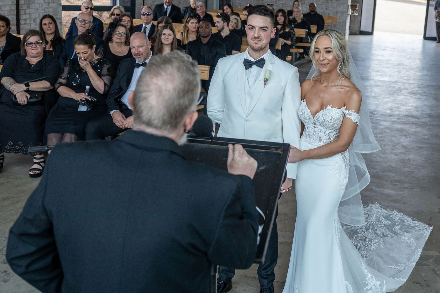 A bride and groom stand before their officiant during the wedding ceremony with guests seated behind them.