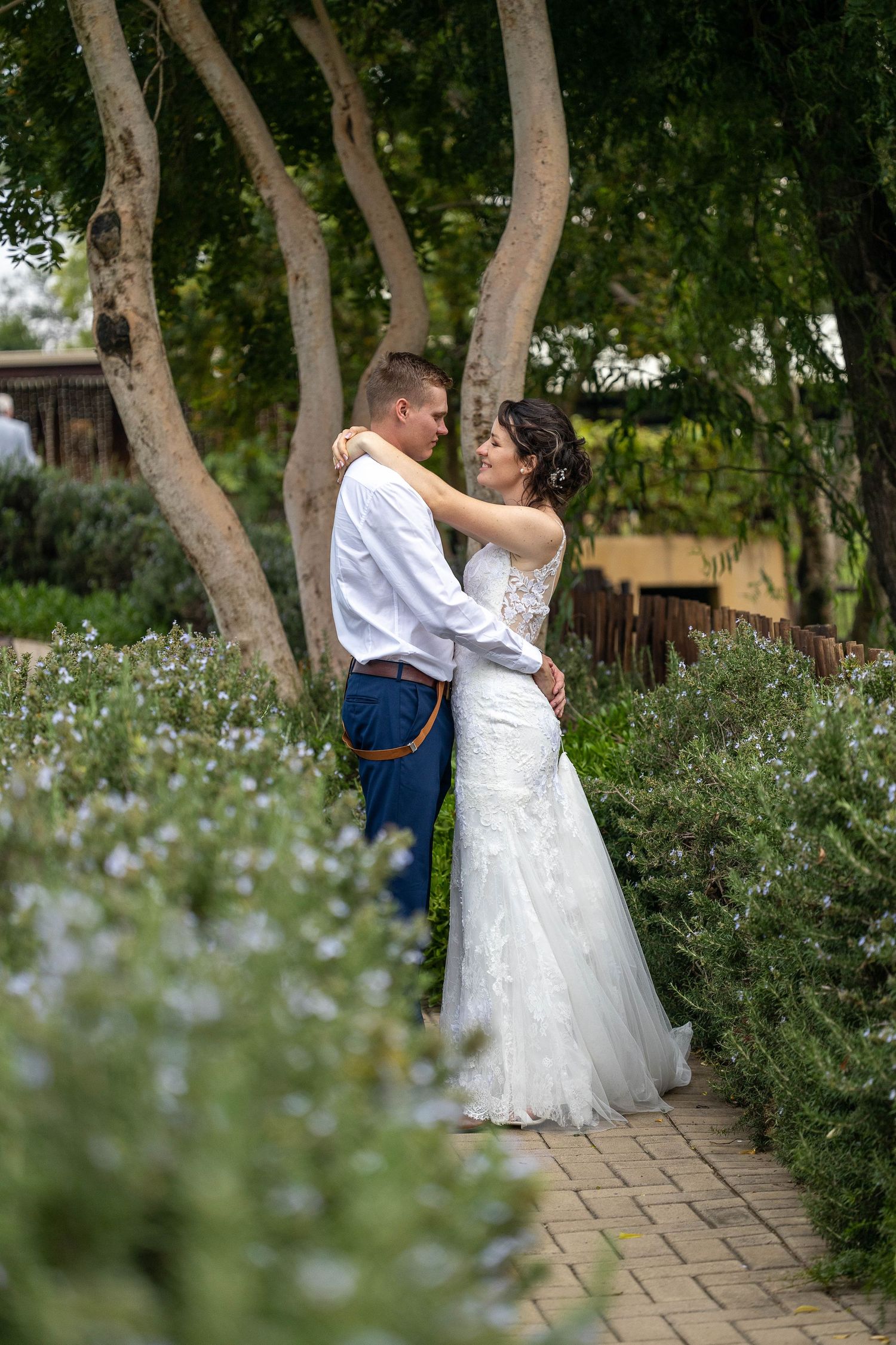 Bride and groom embrace tenderly on a brick pathway surrounded by blooming flowers and tall trees in a garden setting.