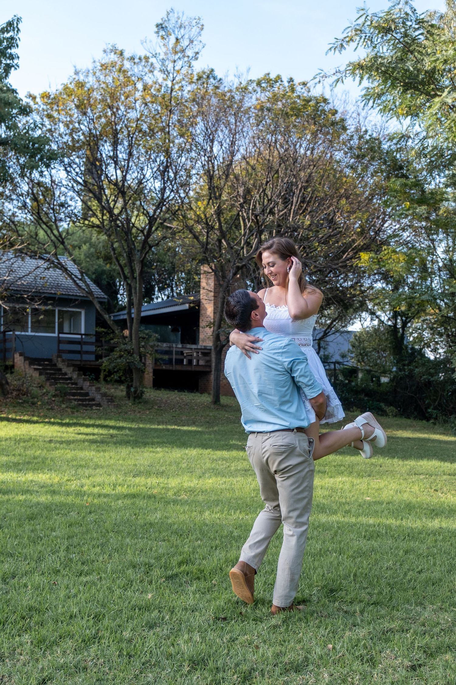 A joyful man lifts a laughing woman in a white dress in a lush green garden.