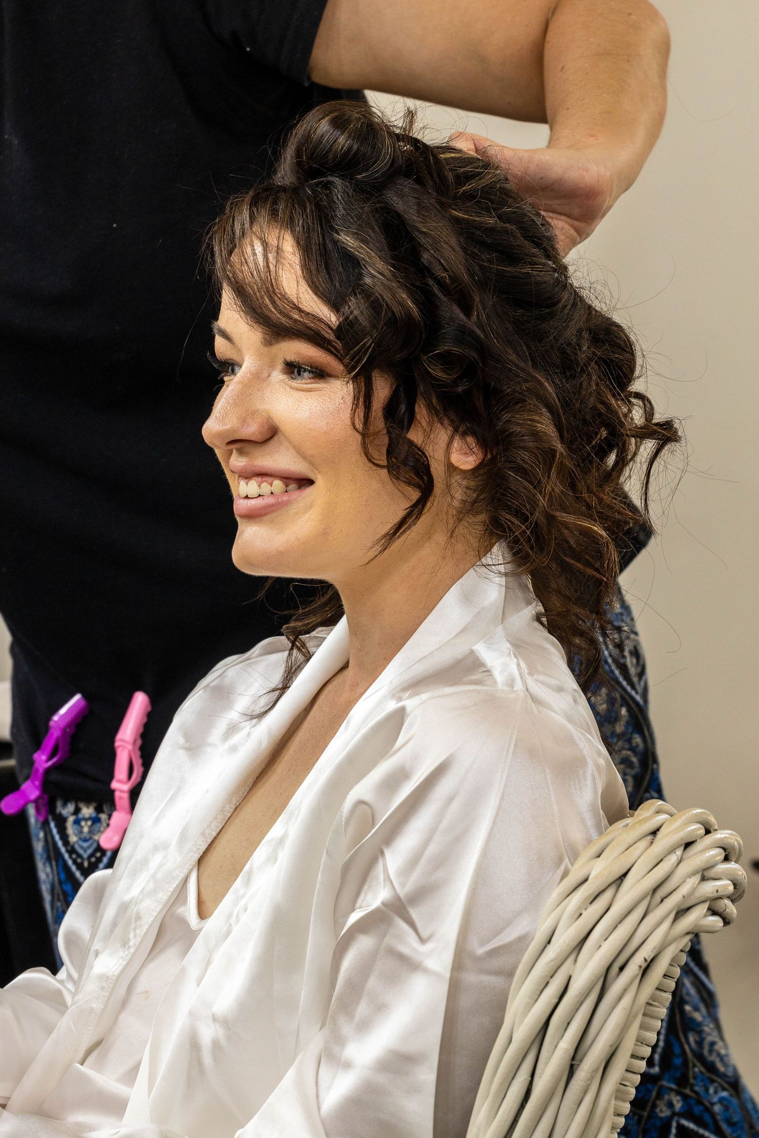 Smiling bride getting her curly brunette hair styled by a hairdresser while wearing a white satin robe.