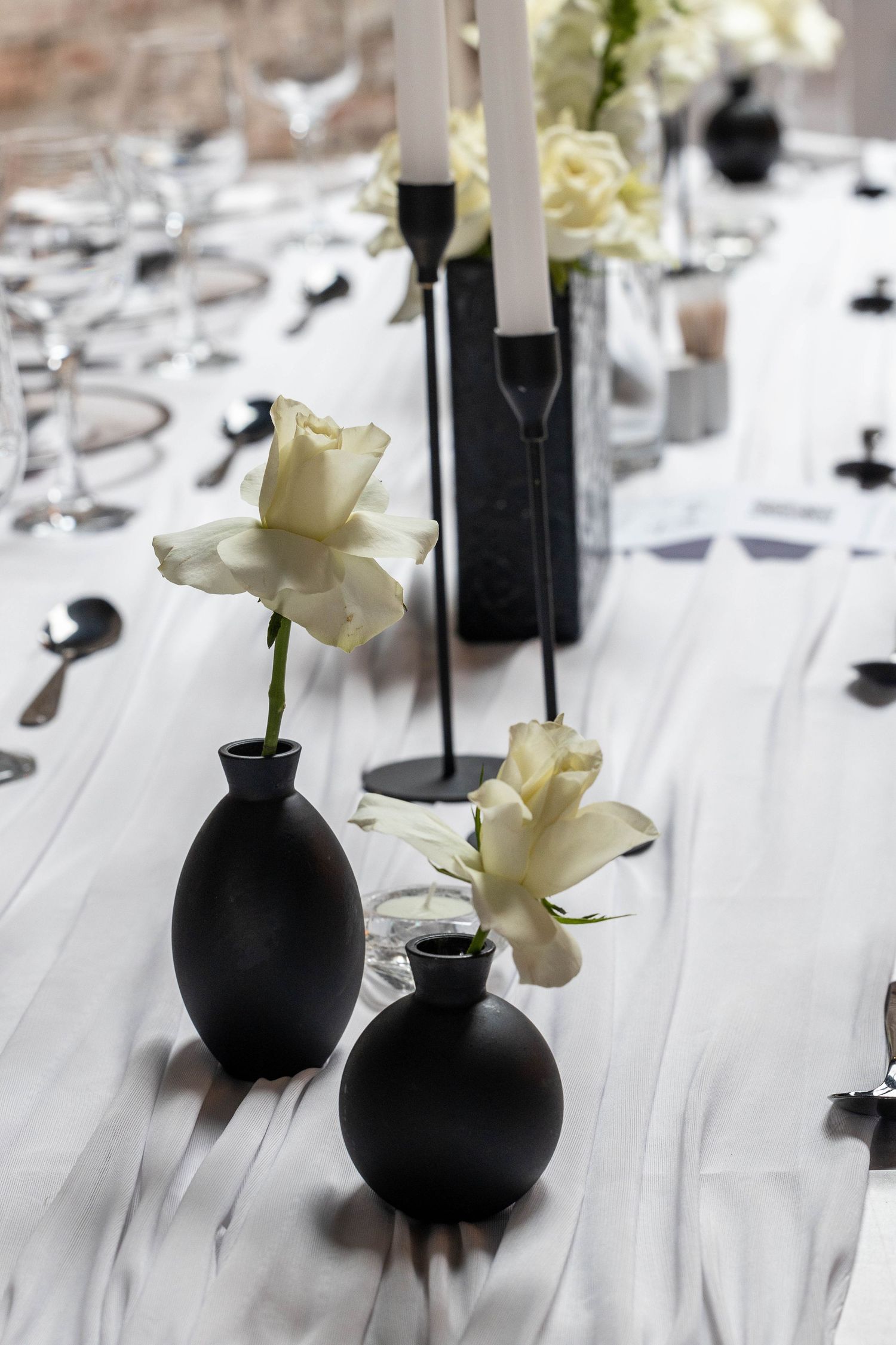 Black matte vases with white roses and candle holders on white marble wedding reception table.