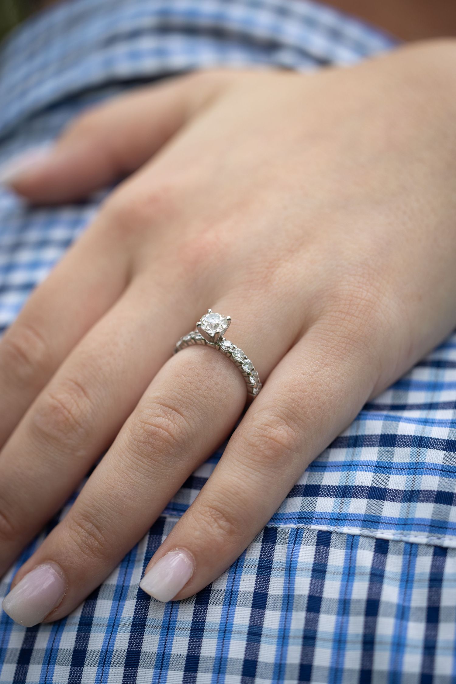 A close-up of a womans hand wearing a delicate diamond solitaire engagement ring on a blue shirt.