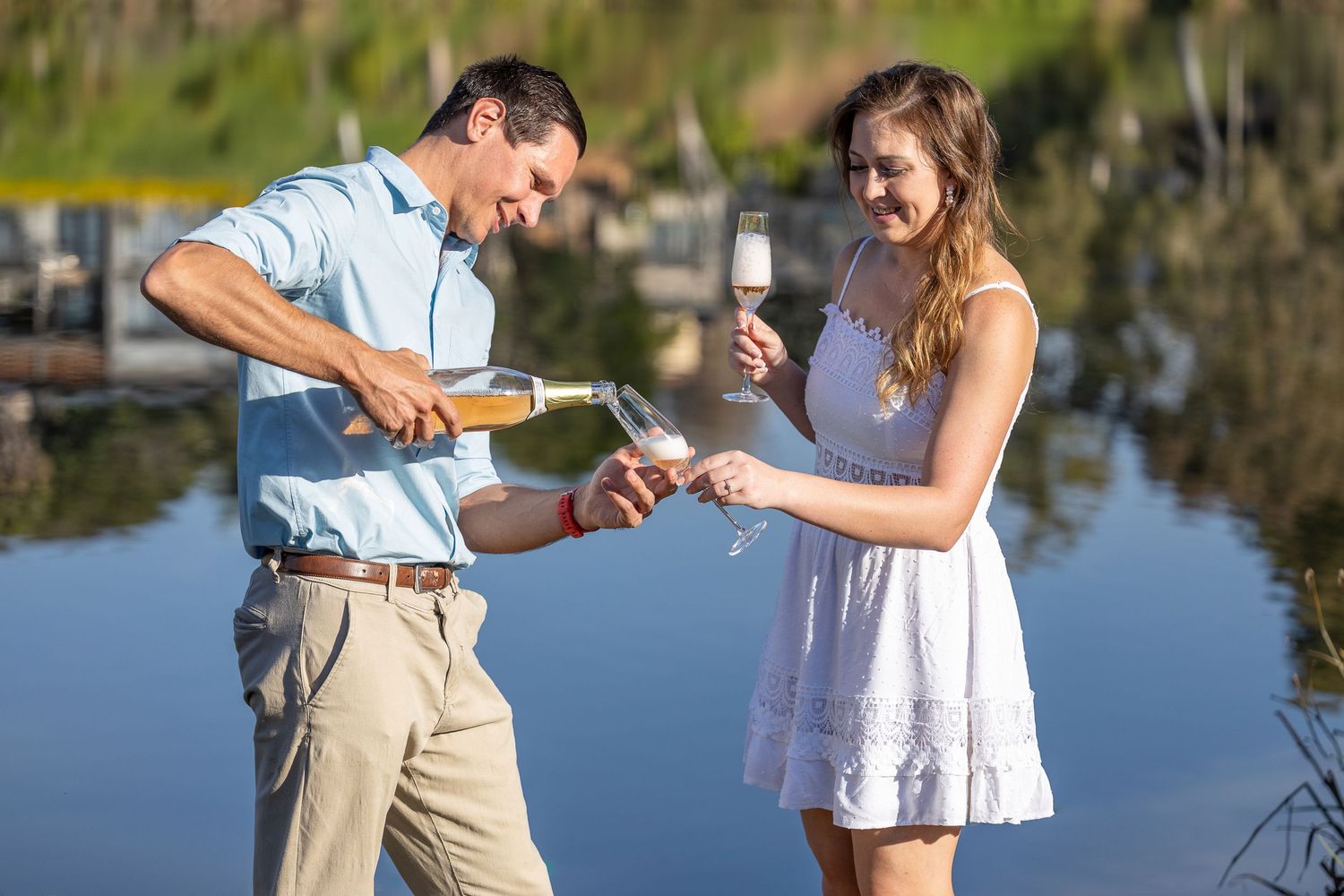A man pours champagne for a woman outdoors near a tranquil waterfront.
