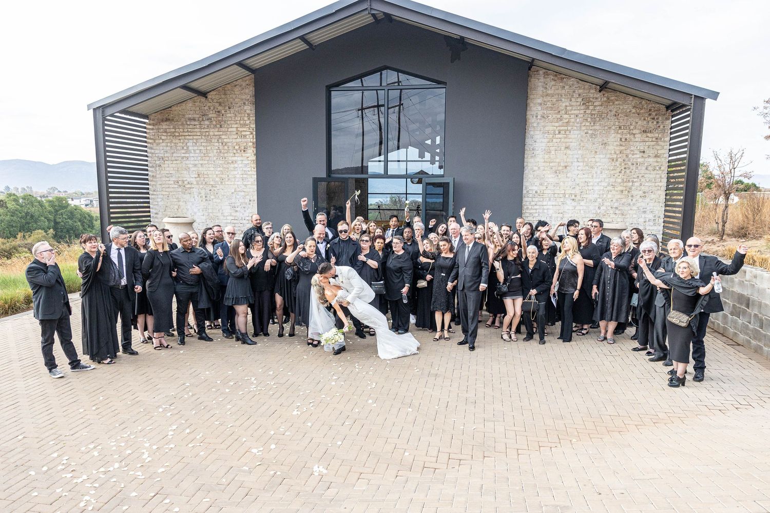 Large wedding party dressed in black poses outside modern stone venue with bride and groom in white at center.