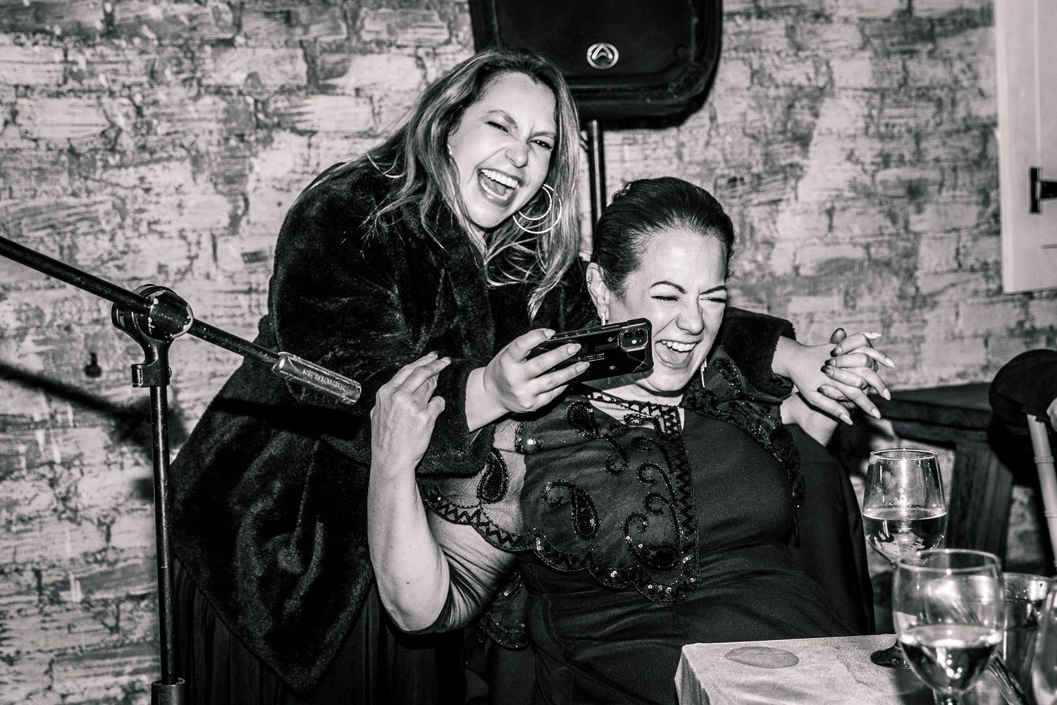 Black and white photo of two women laughing while looking at a phone at a wedding.