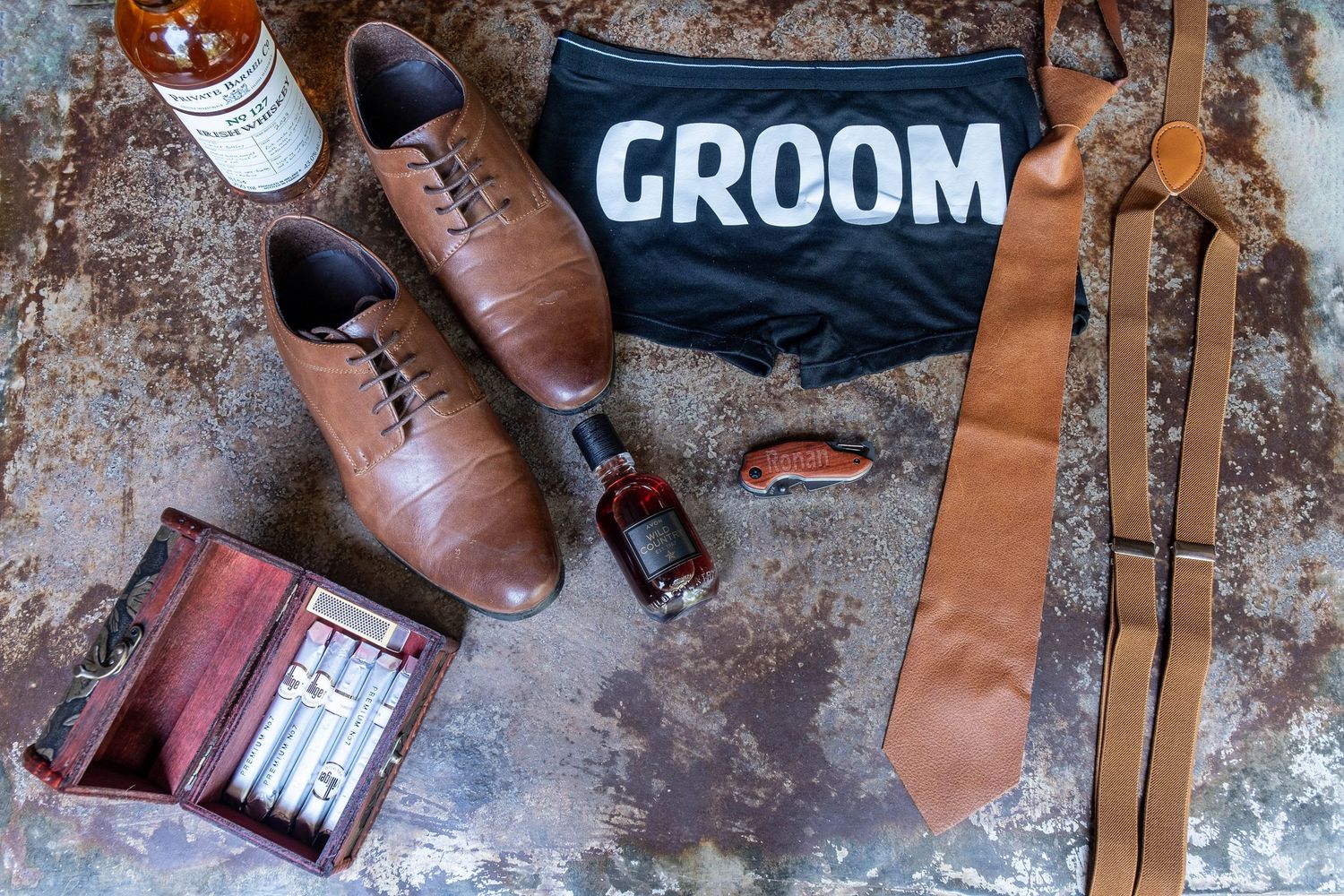 Groom wedding accessories flat lay with brown leather shoes, tie, suspenders, cologne, wallet, and a groom bag.