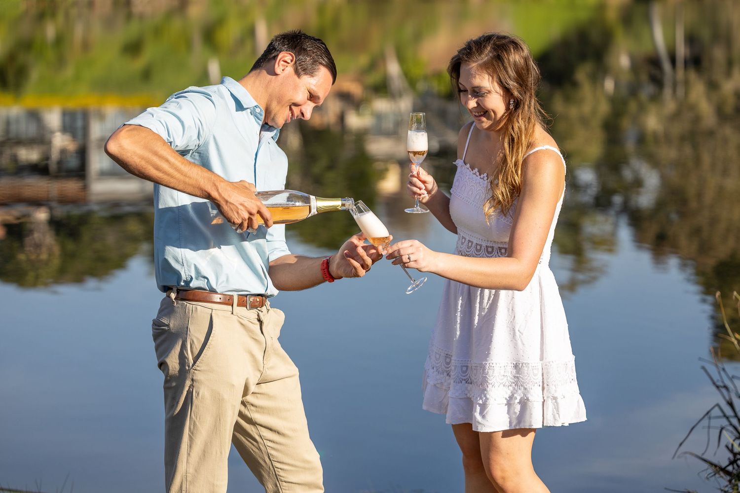 Man pouring champagne for a woman in a white dress beside a shimmering lake at sunset.
