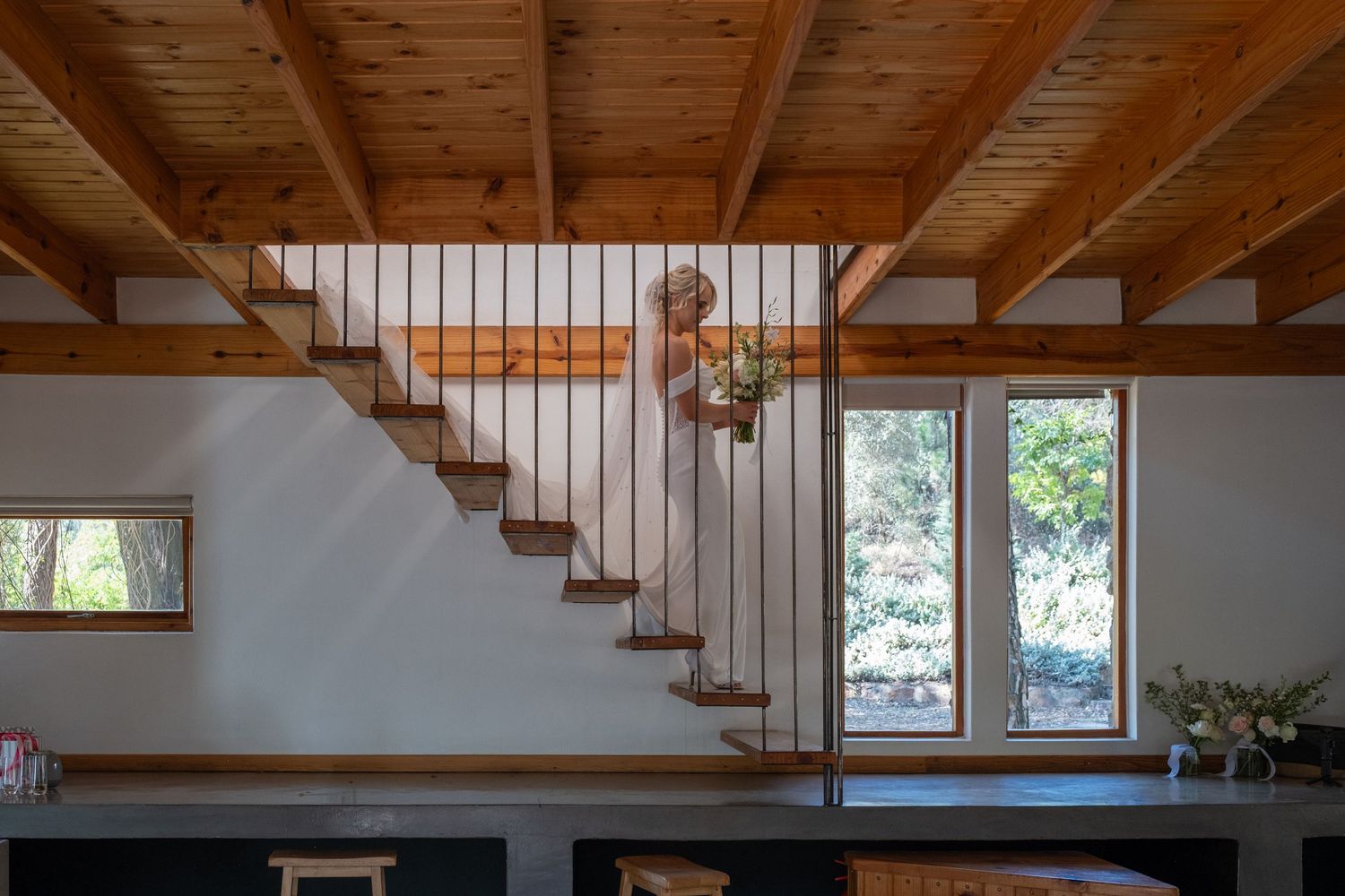 Bride in white gown posing on wooden staircase inside a rustic timber-framed home with natural window light.