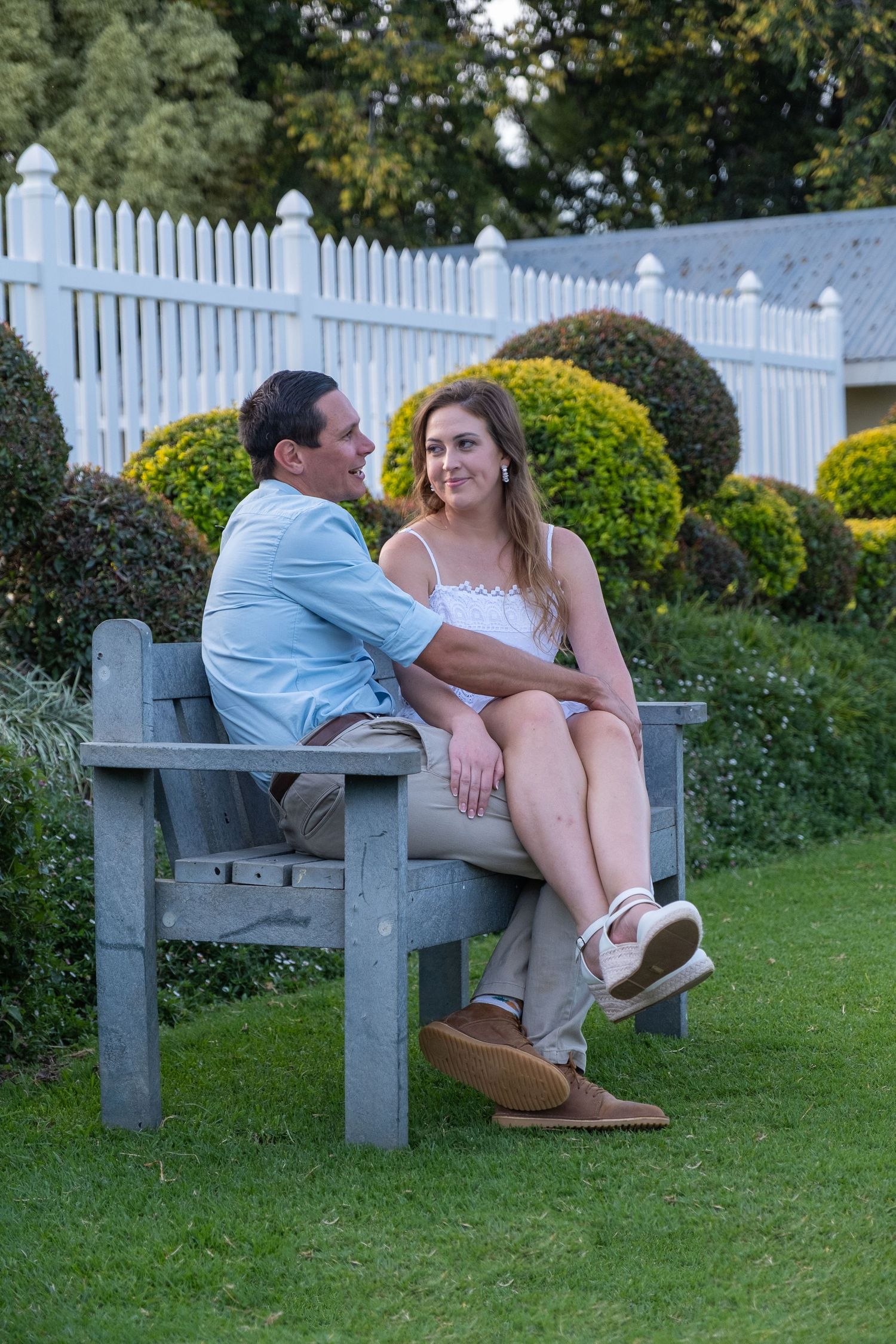 Happy couple sitting together on a wooden garden bench surrounded by manicured hedges and white picket fence.