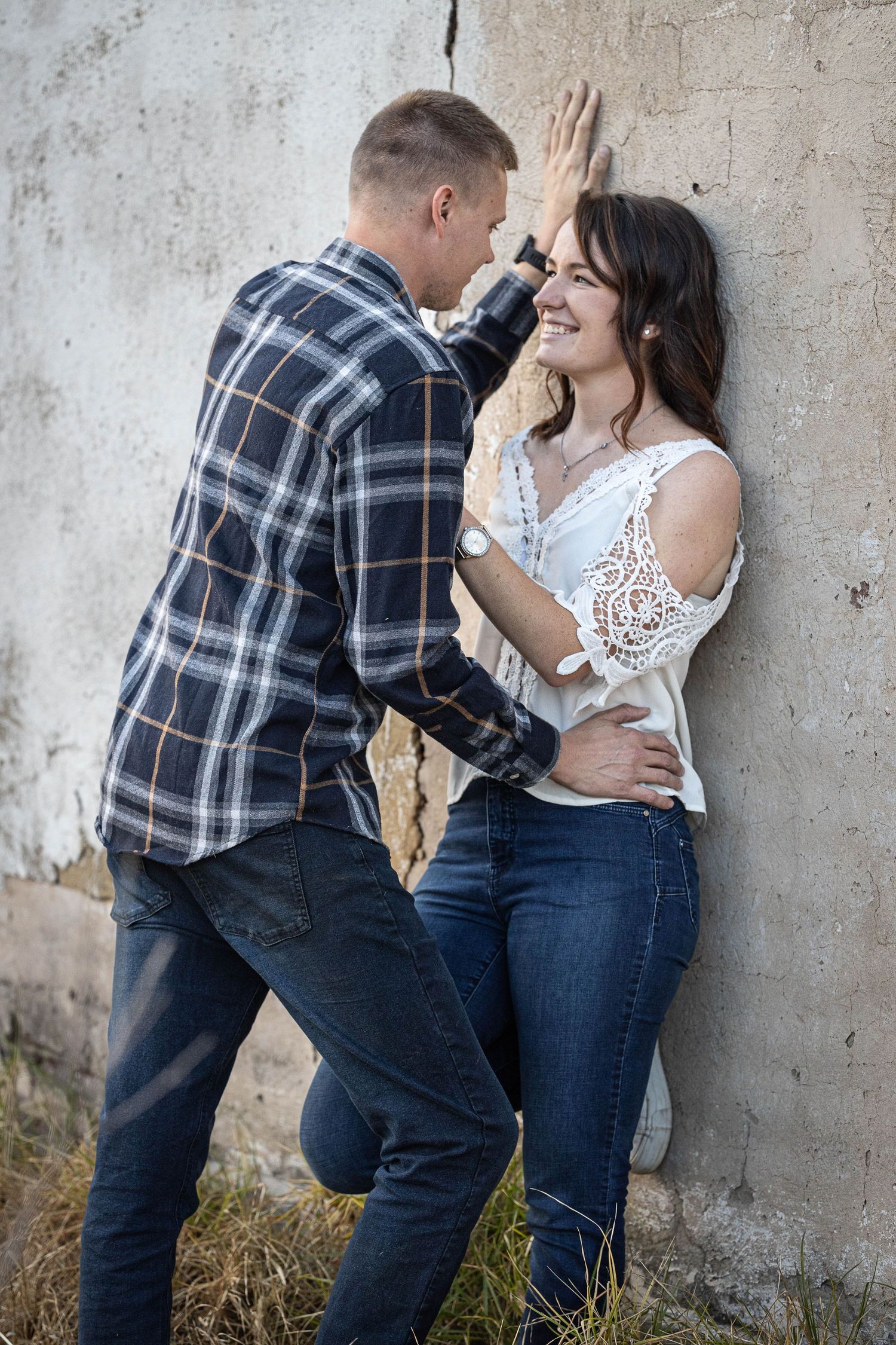 Couple leaning against rustic concrete wall, man placing hand above smiling woman in white lace top and jeans.
