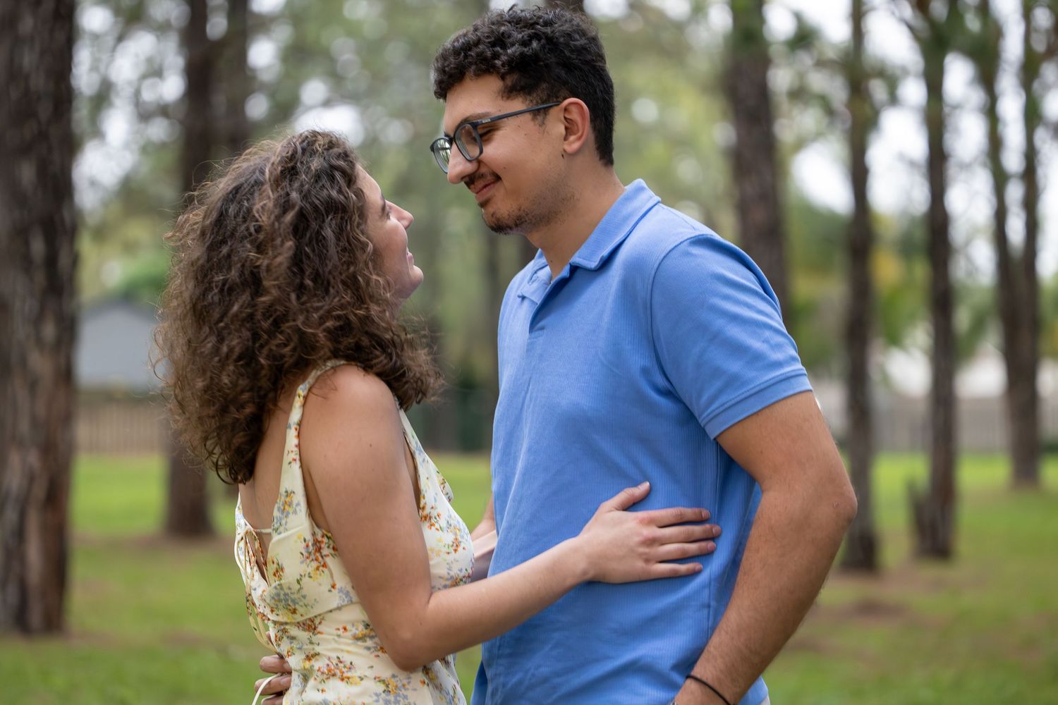 A happy couple embraces outdoors in a park, smiling at each other in casual summer attire.