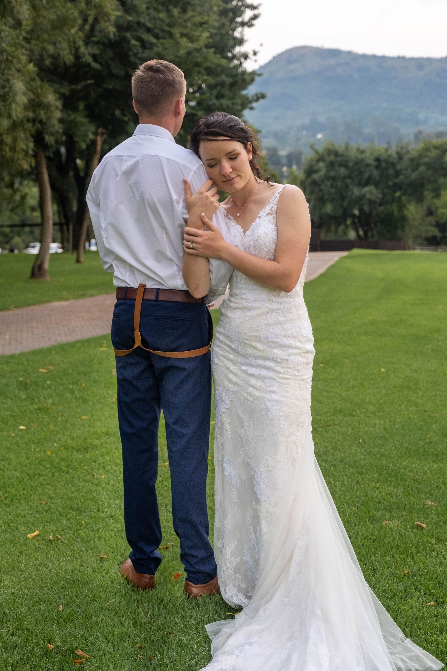 Bride in fitted lace wedding dress embraces groom in white shirt and navy suspenders on a green lawn.