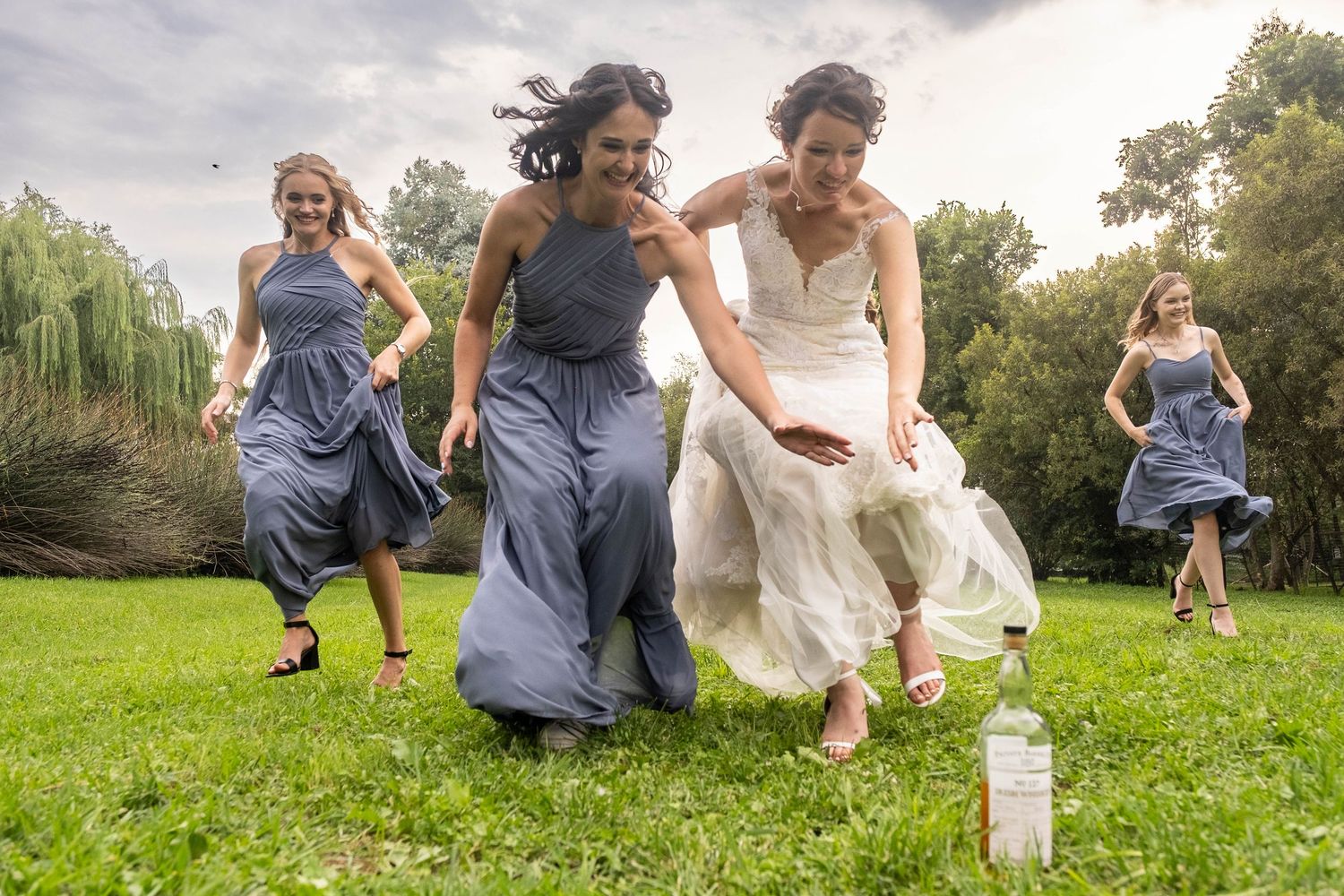 A bride and bridesmaids in blue dresses laugh and run across a green lawn at a wedding.