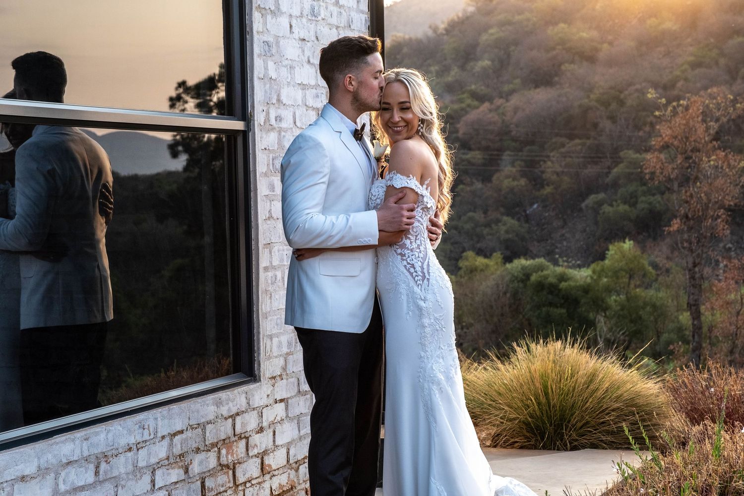 Groom kissing bride on cheek at sunset outside modern white brick wedding venue.