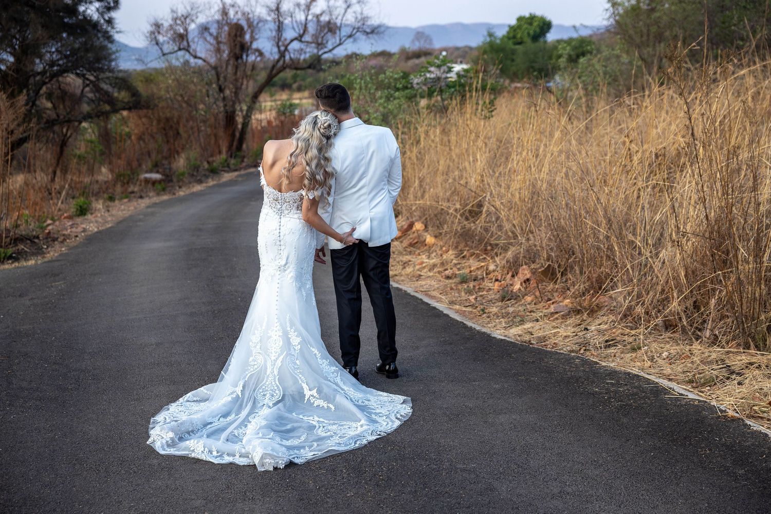 Bride and groom walking hand in hand down a rural road, bride in lace wedding gown with long train.