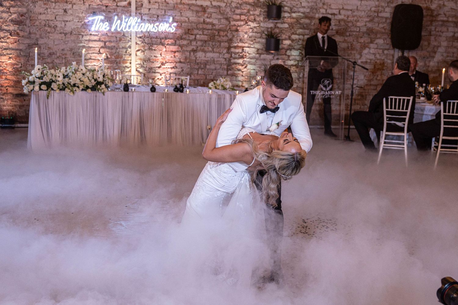 Groom dipping bride dramatically on dance floor with dry ice fog at The Barn wedding venue.