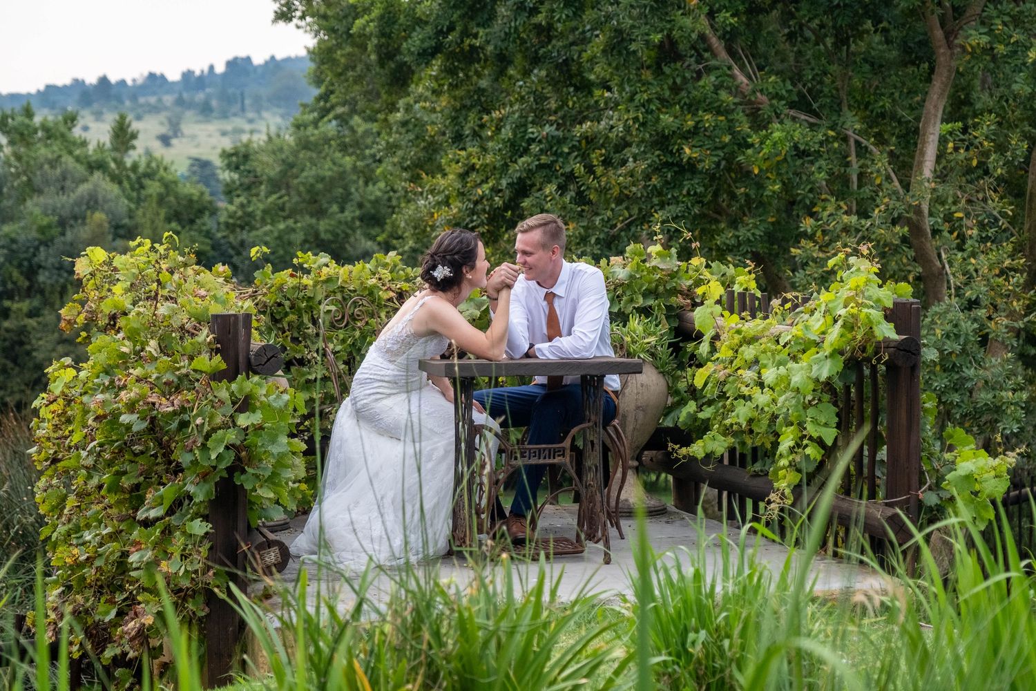 A bride and groom sit together at a small table on a vine-covered terrace surrounded by lush greenery.