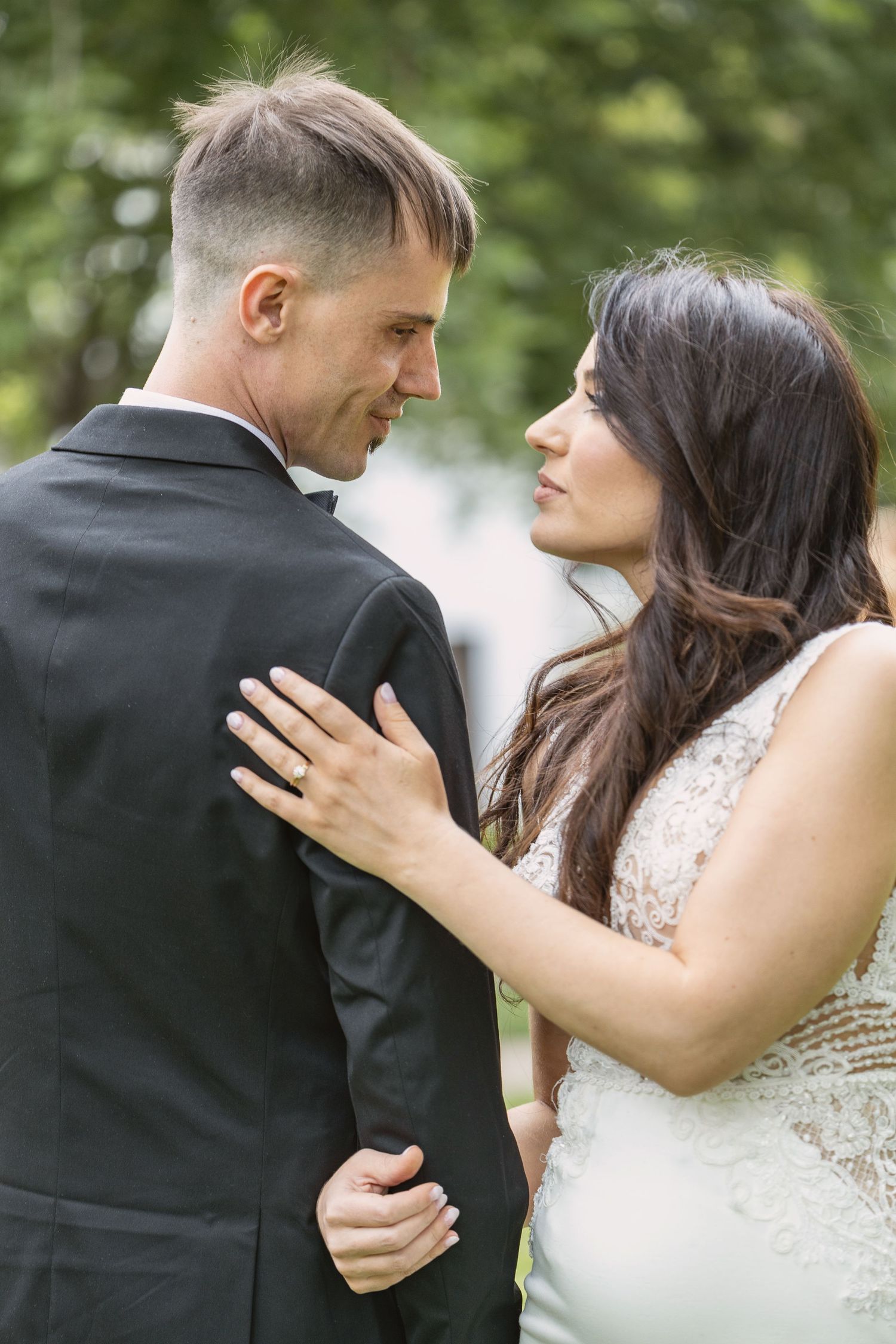 Bride and groom sharing an intimate moment outdoors, bride in lace dress with engagement ring visible.