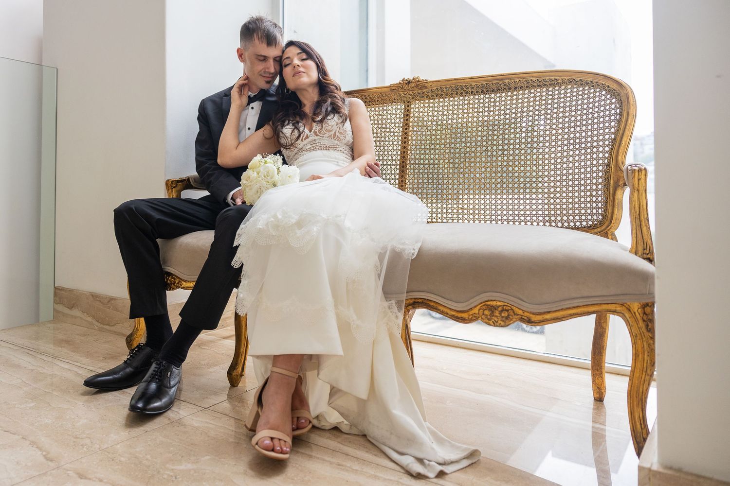 Bride and groom sharing intimate moment seated on elegant gold French-style sofa in bright venue.