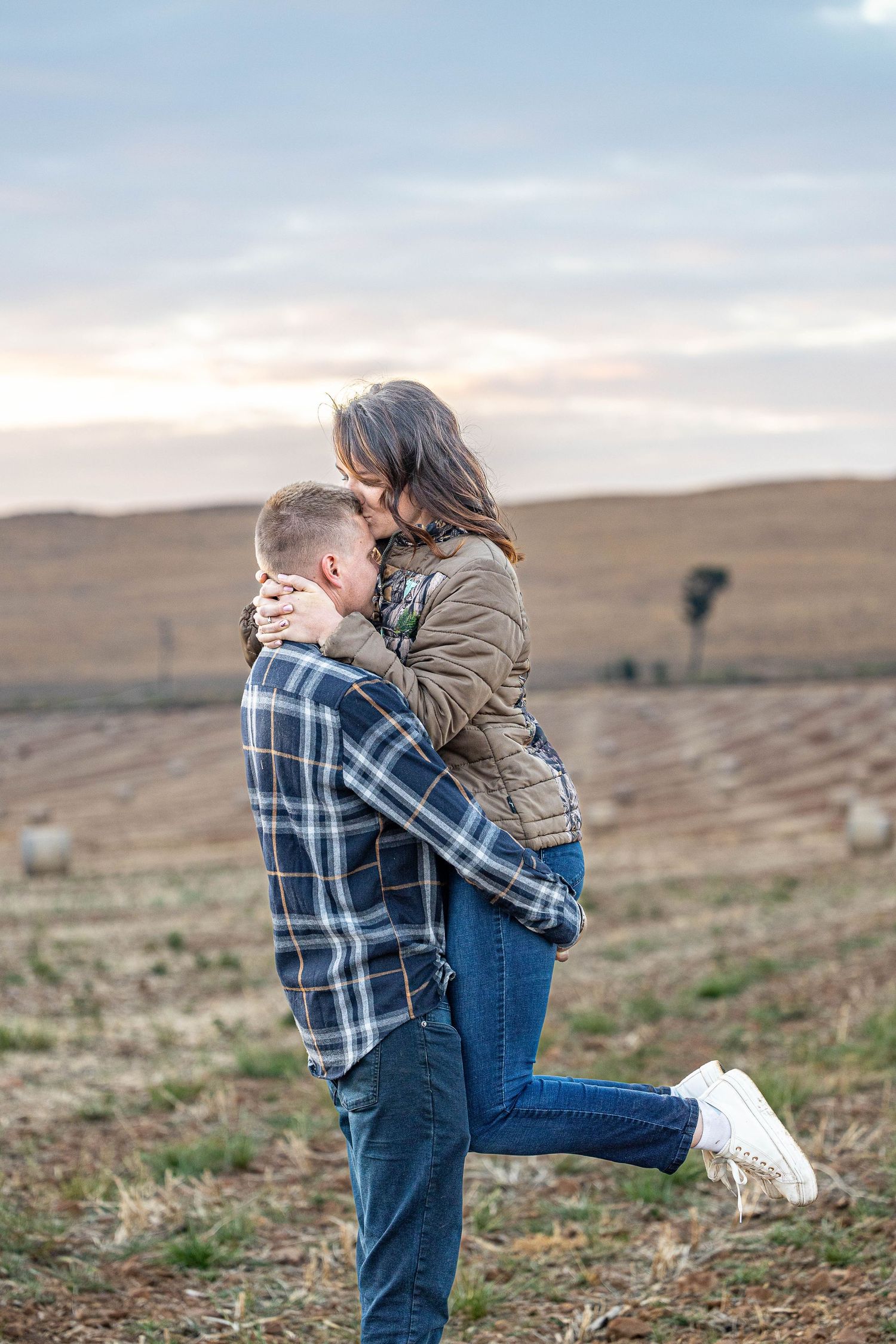 Man lifting woman in tan jacket as they embrace in open harvested field at sunset.