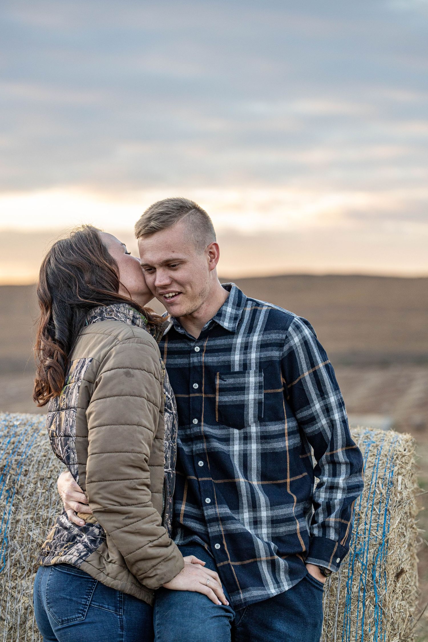 Woman kissing smiling man on cheek while leaning against hay bale in golden field at sunset.