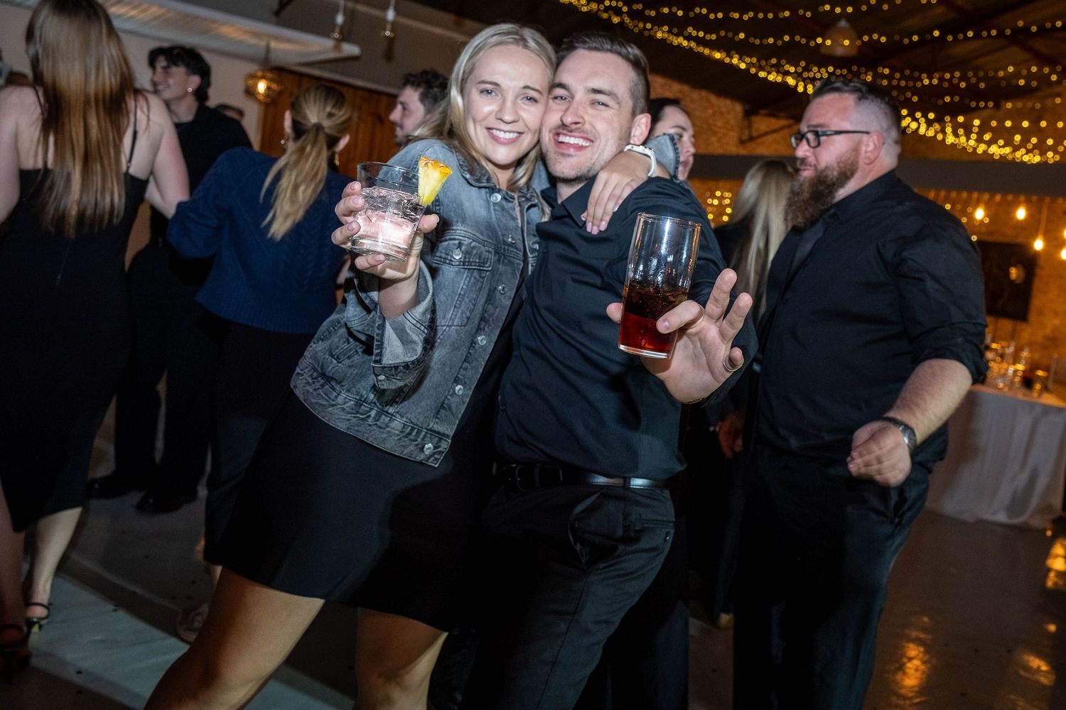 Smiling couple holding wine glasses and posing together at a warmly lit wedding reception.