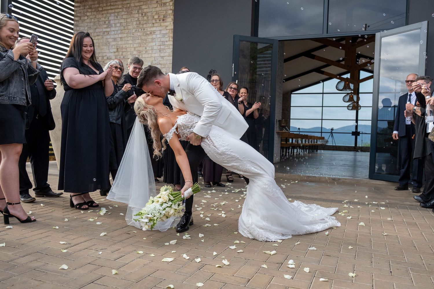 Groom dips bride for romantic kiss as guests celebrate outside wedding venue.